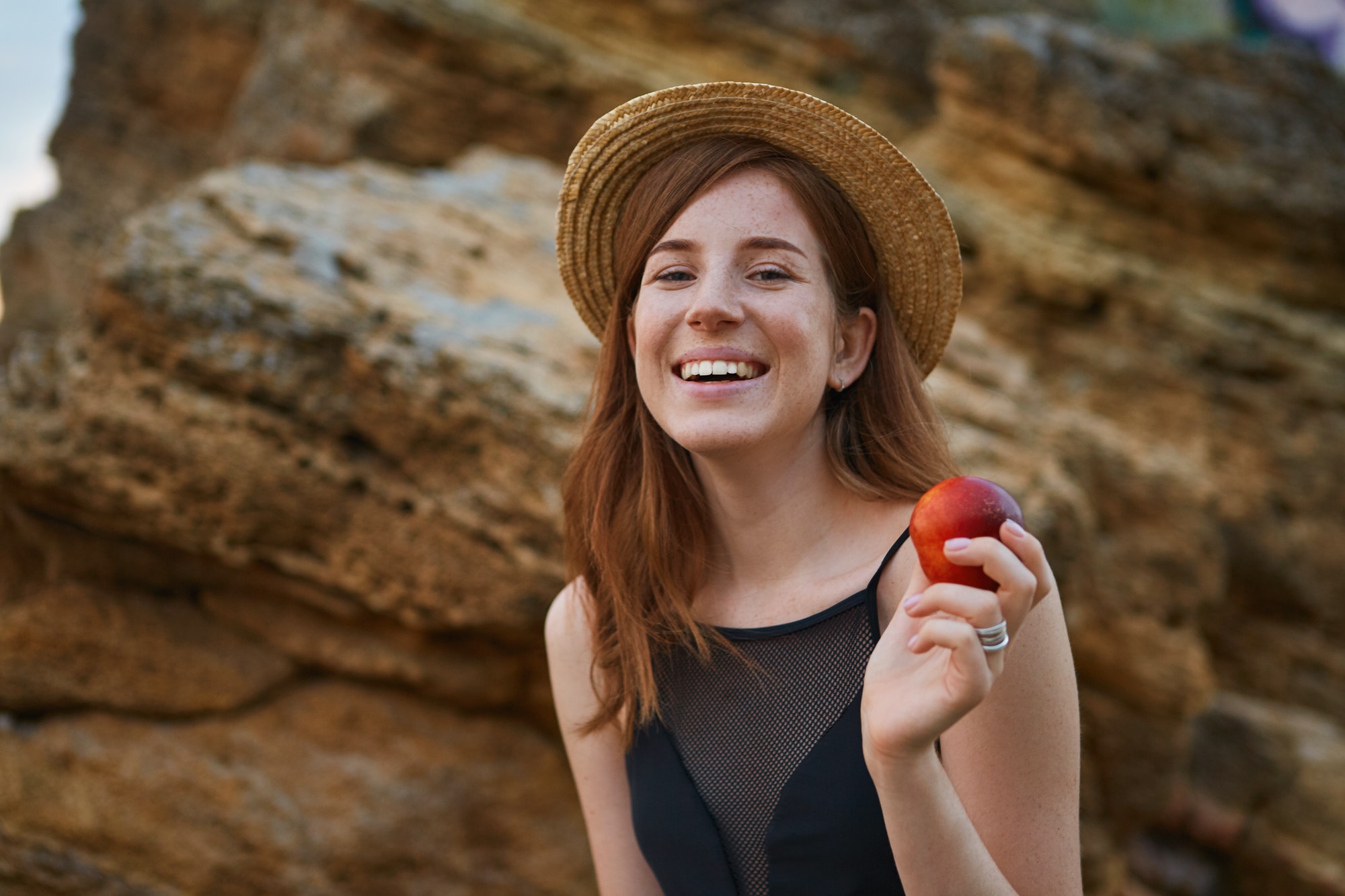 Portrait of young ginger nice freckles lady on the beach, wears hat, eating a peach, broadly smiles